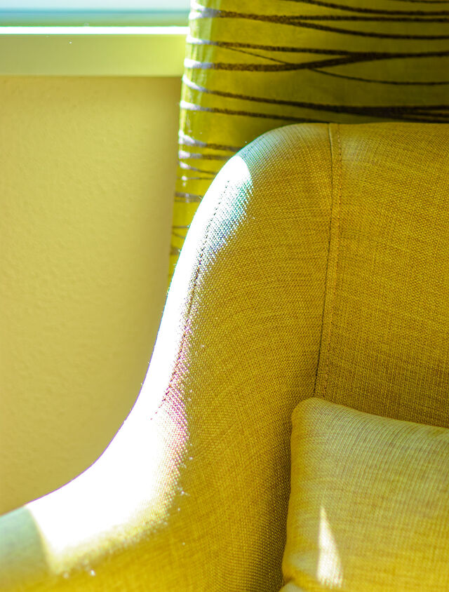 Close-up of warm yellow upholstered reading chair with curved backrest in bright sunlight with metallic green striped Zimmer & Rohde curtains in background in bedroom of furnished apartment Bamberg