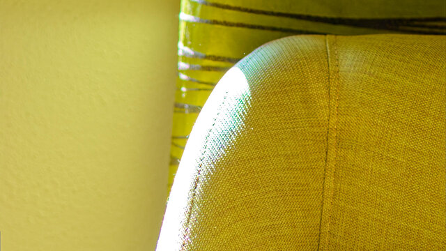 Close-up of warm yellow upholstered reading chair with curved backrest in bright sunlight with metallic green striped Zimmer & Rohde curtains in background in bedroom of furnished apartment Bamberg