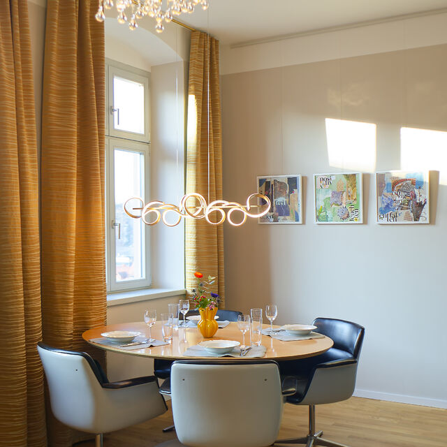 Modern dining area with round table, white and black chairs, gold chandelier, and floor-length curtains in warm tones.