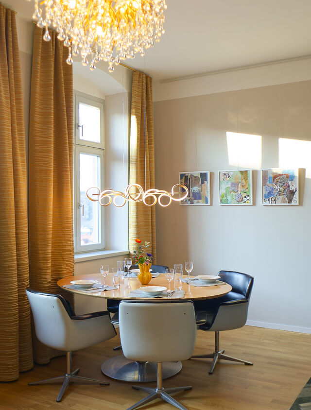 Modern dining area with round table, white and black chairs, gold chandelier, and floor-length curtains in warm tones.