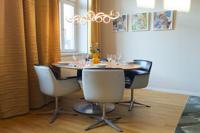 Modern dining area with round table, six white chairs, gold curtains and three wall paintings in bright room with wooden floor.