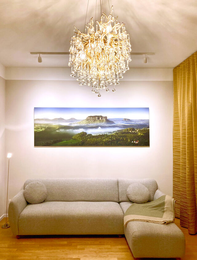 Evening view of living room with gray salt-and-pepper sofa, large-format landscape photograph Lilienstein III by Dirk Wiedlein, crystal Icicle chandelier by Paul Neuhaus and golden Zimmer & Rohde curtains in furnished apartment Bamberg