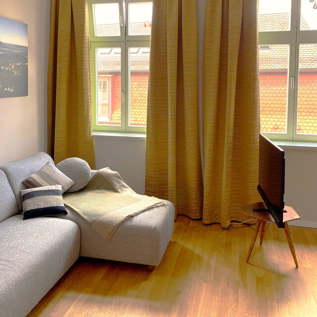 Sunny living room with gray salt-and-pepper sofa, decorative cushions, 45-inch Metz HD television on wooden retro stand, Paul Neuhaus Icicle chandelier, golden Zimmer & Rohde curtains and view of red tile roofs in furnished apartment Bamberg