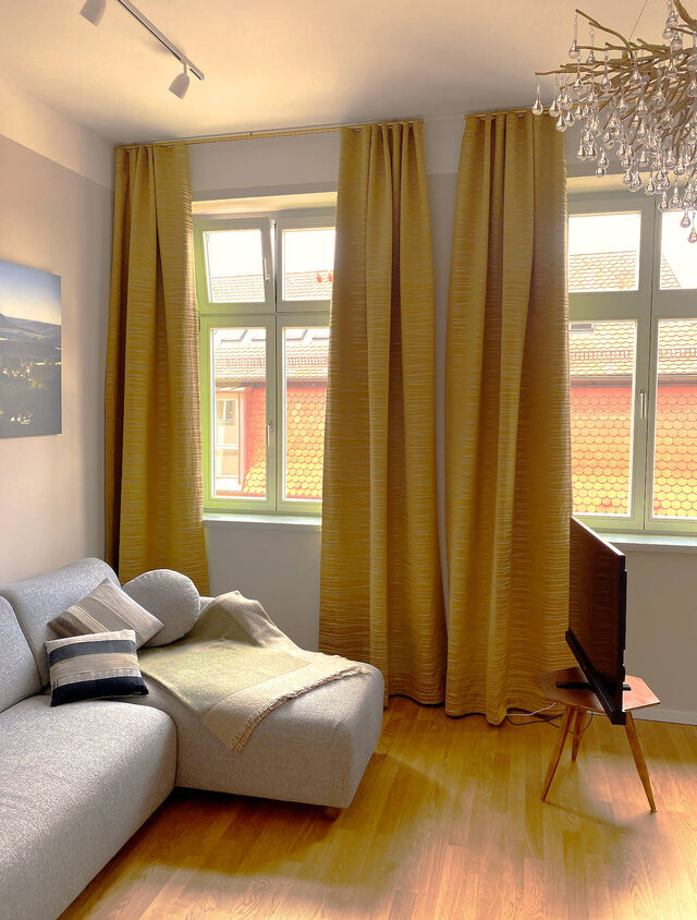 Sunny living room with gray salt-and-pepper sofa, decorative cushions, 45-inch Metz HD television on wooden retro stand, Paul Neuhaus Icicle chandelier, golden Zimmer & Rohde curtains and view of red tile roofs in furnished apartment Bamberg
