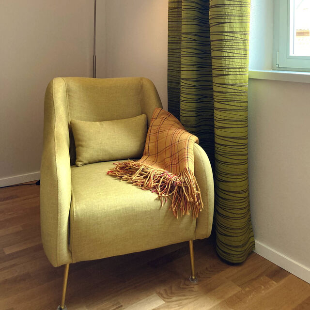 Cozy yellow upholstered armchair with cushion and plaid throw blanket under warm floor lamp next to window with green-yellow striped curtains on oak parquet in bedroom of furnished apartment Bamberg