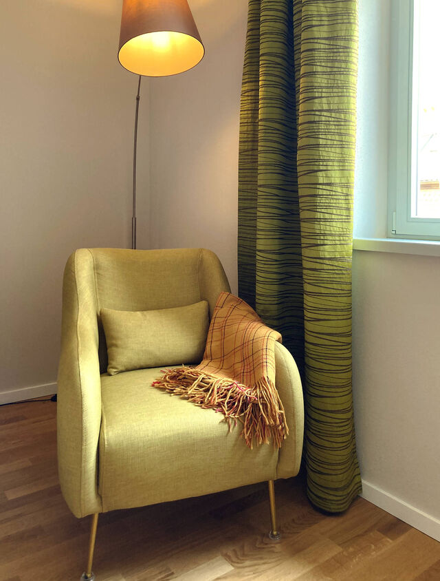 Cozy yellow upholstered armchair with cushion and plaid throw blanket under warm floor lamp next to window with green-yellow striped curtains on oak parquet in bedroom of furnished apartment Bamberg